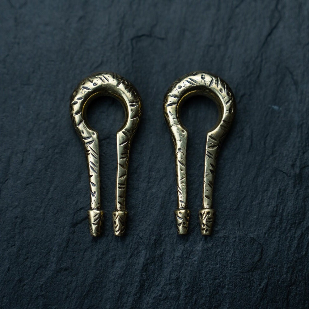 A pair of intricately designed keyhole and hanger-style gold ear weights from the Kaya Movement collection is showcased against a dark textured background. Ideal for the stretched ear game, these minimalist earrings feature engraved patterns and boast an antique appearance.