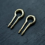 A pair of intricate brass jaw harps with engraved patterns, reminiscent of Kaya Movement's Gold Ear Weights in Keyhole & Hanger Style, rest on a dark, textured surface. The instruments feature a looped design with a narrow, elongated, and slightly curved end.
