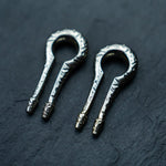 Close-up of two intricately designed Silver White Brass Ear Weights from Kaya Movement, featuring detailed carvings on their circular frames and prongs. Placed side by side on a dark slate surface, these minimalist earrings for stretched lobes resemble antique body mod jewelry with their exquisite craftsmanship and textured, worn appearance.