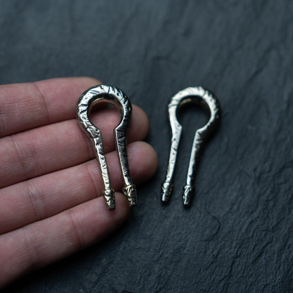 Close-up of a hand holding a small metal musical instrument resembling a mouth harp or jew's harp, with another identical instrument placed on a dark surface next to it. The instruments have a horseshoe shape and appear to be made of textured metal, much like the finely crafted Silver White Brass Ear Weights by Kaya Movement, known for their Keyhole & Hanger Style and minimalist design suitable for stretched lobes.