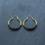The Handmade Boho and Tribal Horn Earrings by Kaya Movement, crafted from natural buffalo horn, feature ornate gold-toned detailing on the upper part and intricately carved black sections on the lower part. These cool, unique statement earrings are ethically sourced and beautifully displayed on a dark, textured surface.