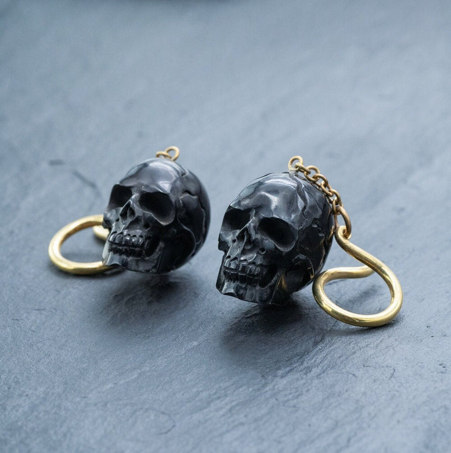 A pair of Handmade Black Skull Dangle Earrings - Gothic Tribal Jewelry for Statement Style by Kaya Movement. These earrings boast detailed hollow eye sockets and teeth, with a glossy finish, attached to gold-colored hooks and chains. They rest on a dark gray flat surface.