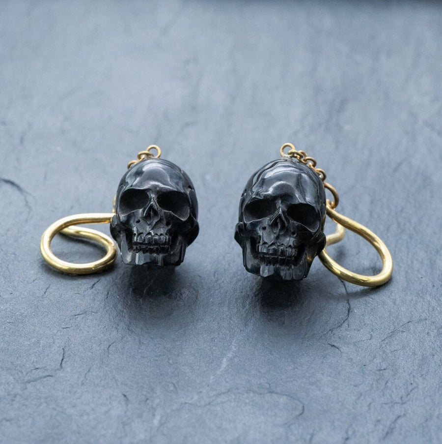 The Handmade Black Skull Dangle Earrings by Kaya Movement, featuring intricate designs and gothic tribal style, are displayed on a textured gray surface. Each earring hangs from a looped gold-colored hook, adding a striking statement to the detailed skull jewelry.