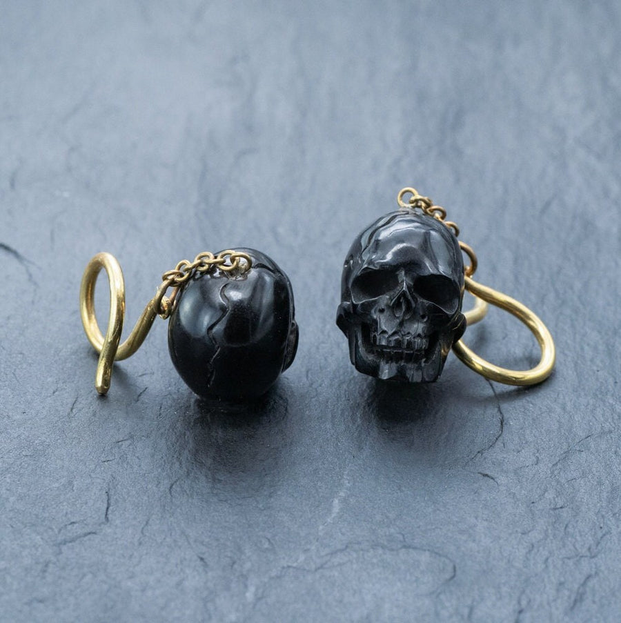 A pair of Kaya Movement's Handmade Black Skull Dangle Earrings, featuring intricate detailing on the black skulls and enhanced by golden hooks and chains, is set against a dark textured surface. The gold components strikingly complement the hauntingly beautiful gothic tribal jewelry, making a distinct statement.