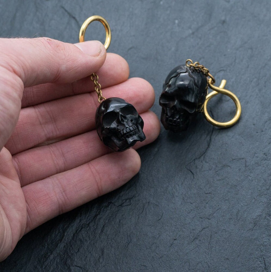A hand holding a keychain featuring a small, black skull with another identical keychain lying nearby on a dark, textured surface. The keychains, resembling the Handmade Black Skull Dangle Earrings - Gothic Tribal Jewelry for Statement Style from Kaya Movement, have gold-colored loops and chains attached to the skulls.