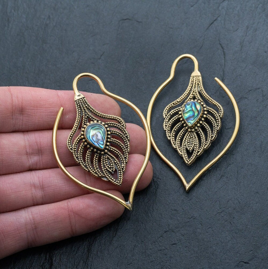Close-up image of a hand holding ornate Kaya Movement Unisex Handmade Brass Leaf Ear Weights with Abalone Shell, designed in the shape of peacock feathers and adorned with intricate detailing. These Boho Hippie Tribal Earrings feature an iridescent gemstone resembling abalone shell and the hand provides a sense of scale for this edgy urban fashion jewelry.