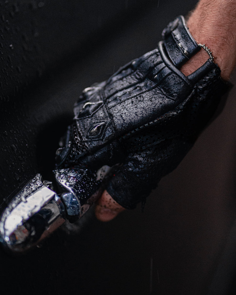 A close-up shot of a hand wearing Kaya Movement's Custom Dragon Scale Leather Gloves—black, textured, and fingerless—gripping a metallic car door handle. The well-worn leather gloves enhance the scene, with raindrops on the car door adding to the dramatic, moody atmosphere—ideal for motorcycle enthusiasts who appreciate post-apocalyptic goth fashion.