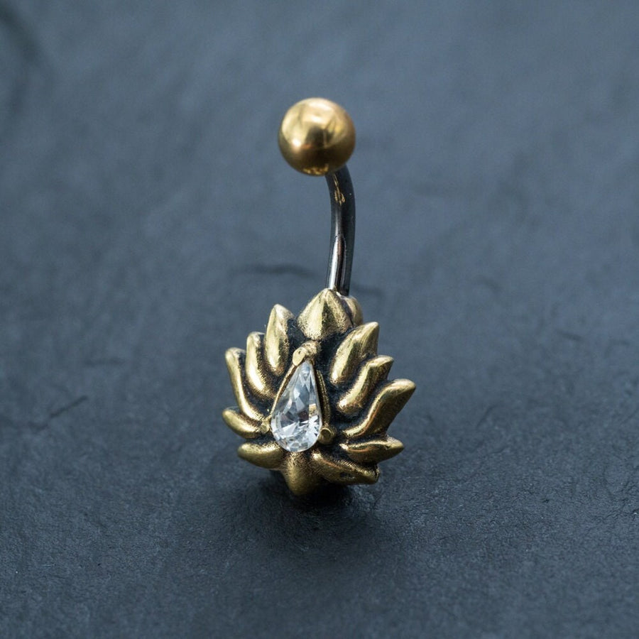 Introducing the Gold Lotus Flower Belly Ring from Kaya Movement. This stunning piece features a teardrop-shaped zircon gemstone elegantly surrounded by lotus petals. The 9mm belly bar is complemented with a small, spherical bead at the top, and is displayed beautifully against a dark textured background. Perfect for adding a touch of sophistication to your navel jewelry collection.