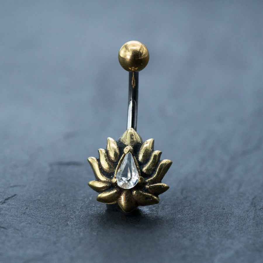 A gold Lotus Flower Belly Ring by Kaya Movement, featuring a zircon gemstone teardrop piercing, is centered on a slate-gray surface. This elegant navel jewelry includes a 9mm belly bar with a gold sphere at the top and a clear zircon gemstone, teardrop-shaped, gracefully hanging from the center of the lotus flower design.