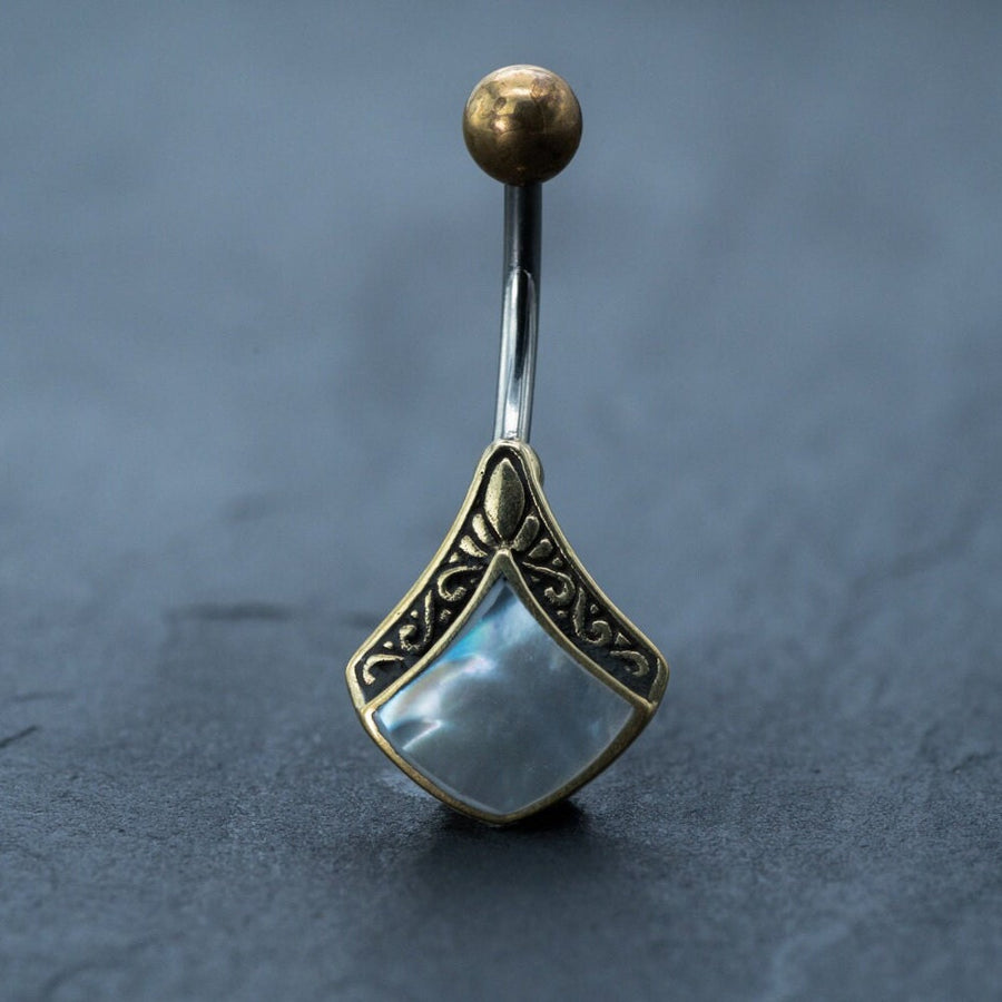 A close-up photo showcasing the Kaya Movement's Mother of Pearl Belly Ring, a boho 9mm navel bar featuring an iridescent, diamond-shaped gem. The intricate designs on the gold setting and the elegant pearl piercing rest on a dark, textured surface.