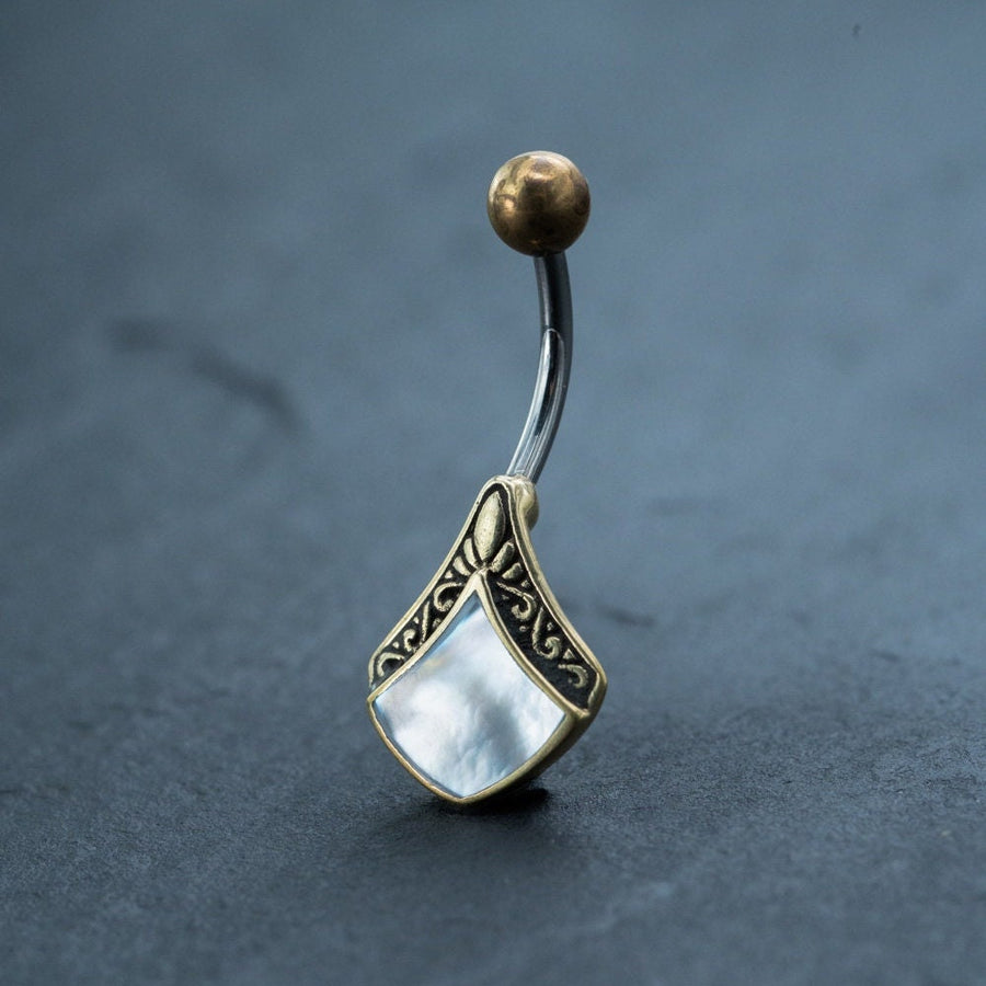 A close-up photo of the Mother of Pearl Belly Ring by Kaya Movement, featuring a curved 9mm metal bananabell bar. One end displays a small, textured gold sphere, while the other end showcases an intricately designed charm with a diamond-shaped mother-of-pearl inlay, creating a Boho vibe against the dark, blurred background.