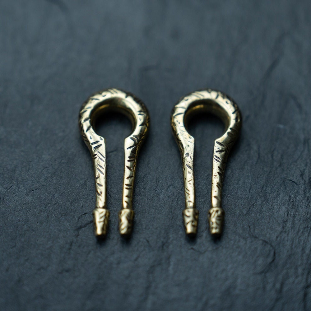 A pair of ornate, metallic objects resembling miniature keys with rounded tops and two prongs at the bottom, resting on a dark, textured surface. These Kaya Movement Gold Ear Weights in Keyhole & Hanger Style feature intricate engravings and could be mistaken for exquisite body mod jewelry designed for stretched lobes.