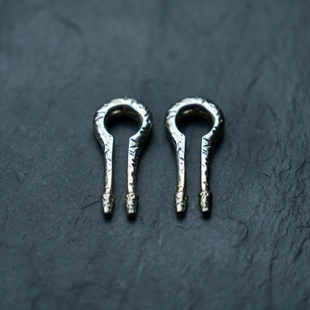 Two shiny metallic harp picks with intricate patterns displayed on a dark textured surface resemble the elegant Silver White Brass Ear Weights from Kaya Movement. The picks are identical and symmetrically positioned side by side, evoking the precision of body mod jewelry designed for stretched lobes.