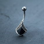 A close-up of the Belly Button Rings Real SeaShell Navel Mother Of Pearl by Kaya Movement showcases a decorative design. The silver 925 belly bar features a curved, 14g surgical steel barbell topped with a small, shiny ball. The bottom of the ring is adorned with a black, shield-like pendant exhibiting intricate ornamental detailing on the edges. This piece is perfect for navel piercings and is available for fast delivery, ideal for adding elegance to summer belly dance attire.