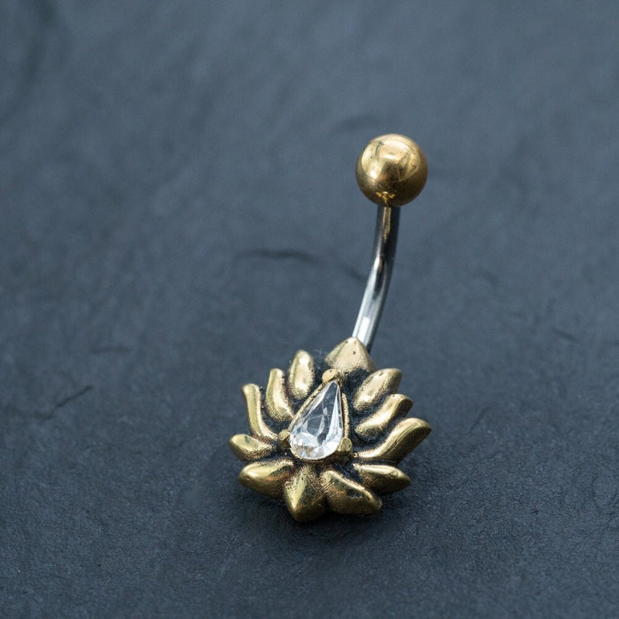 The Kaya Movement Gold Lotus Flower Belly Ring is a stunning piece of navel jewelry featuring a delicate floral design and centered with a teardrop-shaped zircon gemstone. This exquisite 9mm belly bar is beautifully displayed against a dark, textured background.