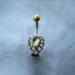 The "Abalone Shell Belly Button Rings" by Kaya Movement, featuring a 9mm Bananabell made from surgical steel, elegantly display an iridescent, teardrop-shaped Abalone Shell center stone. The chic boho bellybutton piercing stands out perfectly on a smooth, dark surface, highlighting the stunning beauty of navel piercings.