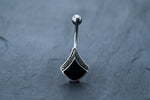 A close-up of a **Black Pearl Belly Rings, Silver Navel Tribal Bars** by **Kaya Movement**, showcasing an intricate, ethnic-inspired design. The jewelry features a metallic silver ball at one end and a shield-shaped piece with a black, ornate pattern at the other, set against a dark, textured background.