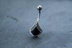 A close-up of a sterling silver body jewelry belly button ring with a curved barbell design. The Kaya Movement Black Pearl Belly Rings feature a teardrop-shaped black gem inlaid in an intricate ethnic-inspired silver setting, resting on a smooth, dark gray surface.