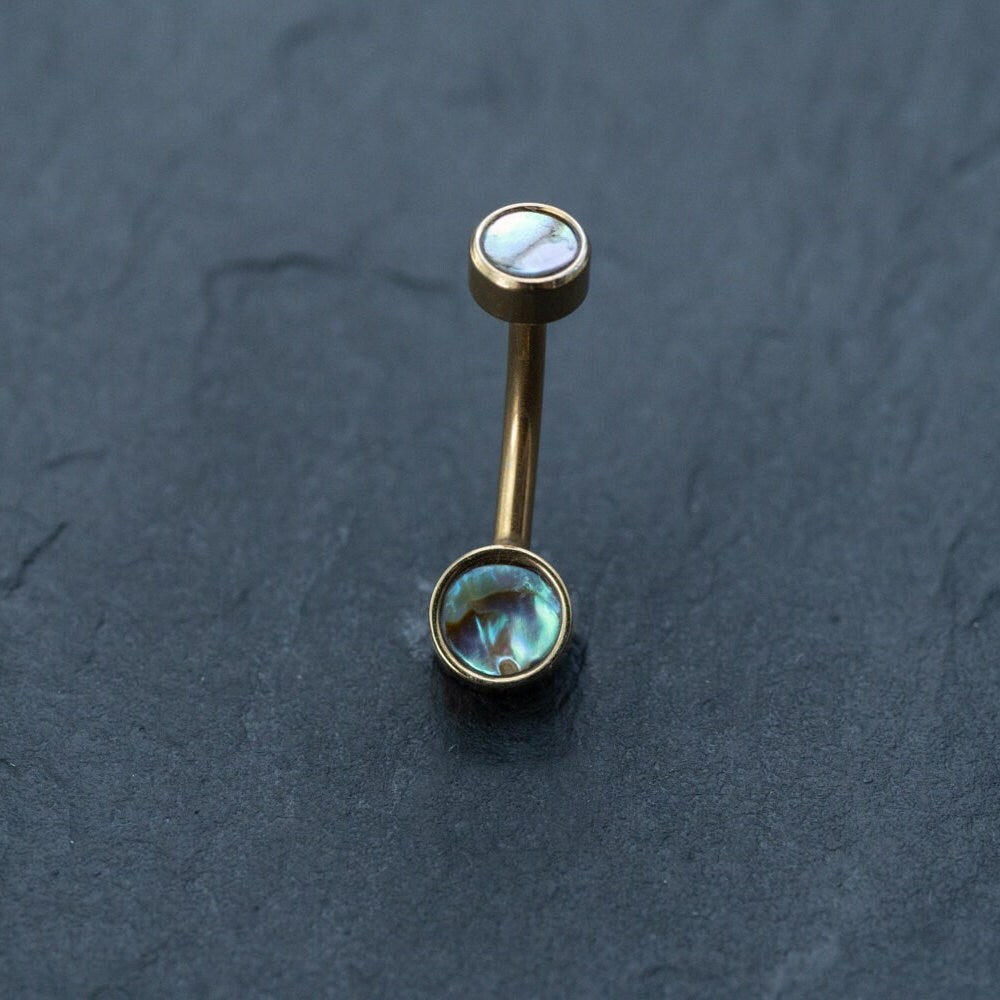 A Kaya Movement gold-toned curved barbell earring, featuring two round ends with shimmering abalone shell inlays, resembles an elegant piece from their boho-chic collection of 18k navel piercing and gold navel rings. This discreet, hypoallergenic belly jewelry is artfully displayed on a dark, textured surface.
