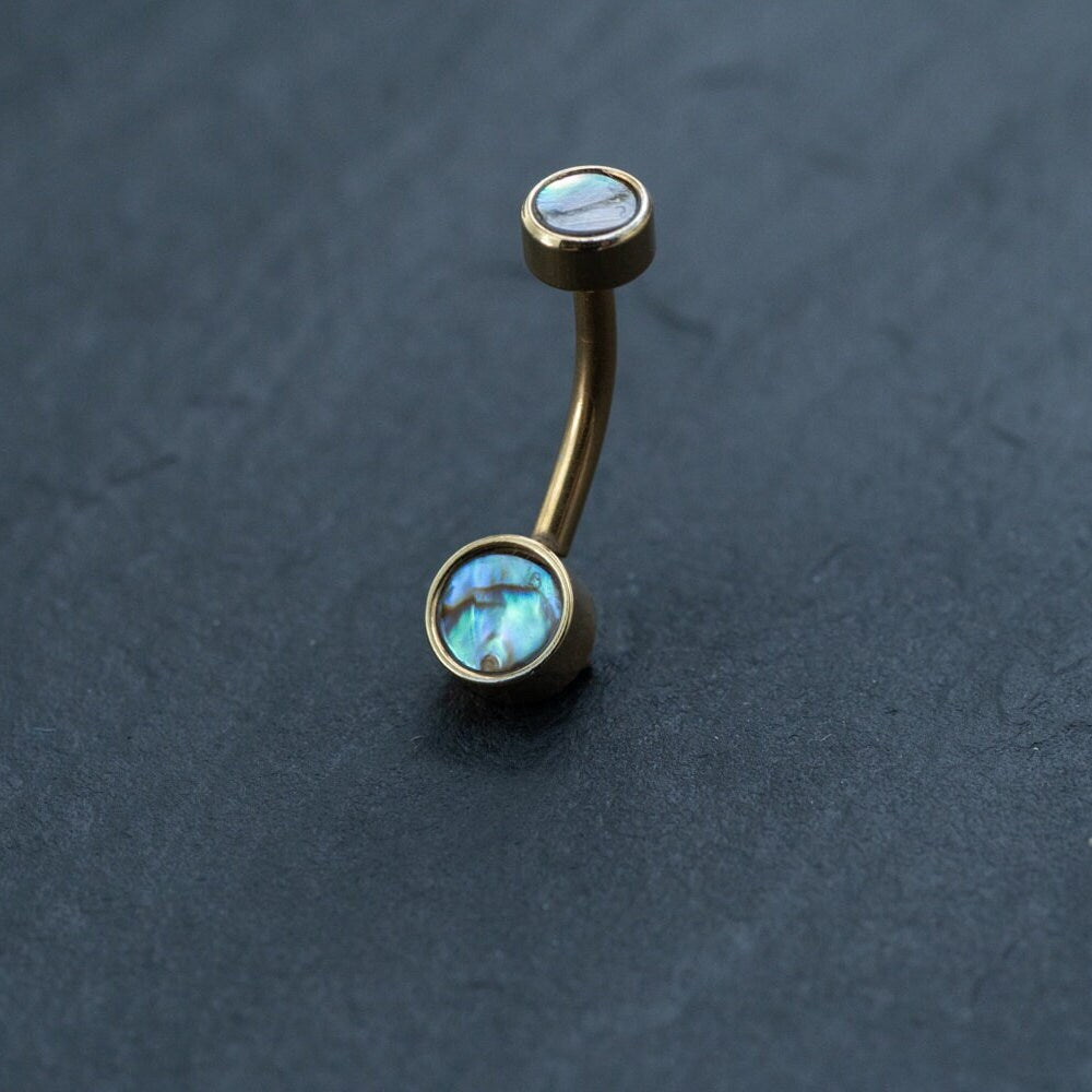 An 18k navel piercing and gold navel ring from Kaya Movement, featuring two abalone shell inlays, evokes the elegance of boho-chic style belly jewelry against a dark, textured background. This hypoallergenic, discreet body jewelry is a perfect piece for those seeking exquisite design.