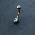 An 18k navel piercing and gold navel ring from Kaya Movement, featuring two abalone shell inlays, evokes the elegance of boho-chic style belly jewelry against a dark, textured background. This hypoallergenic, discreet body jewelry is a perfect piece for those seeking exquisite design.