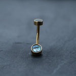 A close-up of Kaya Movement's 18k Navel Piercing & Gold Navel Ring on a dark surface showcases its boho-chic elegance. The hypoallergenic belly jewelry features a barbell design with two ends, each adorned with circular abalone shell inlays. The larger inlay is positioned at the bottom while the smaller one is at the top, enhancing its discreet yet stylish appearance.