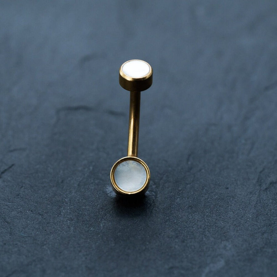 An 18k gold navel ring from Kaya Movement, featuring two white mother-of-pearl circular ends, rests on a dark grey textured surface. The top and bottom ends are similar in size, giving this hypoallergenic, boho-chic belly jewelry a balanced and symmetrical appearance.