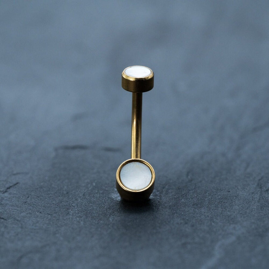 An 18k gold navel ring from Kaya Movement, featuring boho-chic, hypoallergenic MOP shell stones at each end, stands upright on a smooth, dark blue-gray surface. The sleek and polished appearance of this discreet body jewelry is highlighted against the contrasting background.