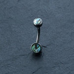 The Kaya Movement Belly Button Jewelry Hypoallergenic Navel Ring, featuring shimmering blue-green abalone shell inlays on both ends and made from sleek Surgical Steel 316L, is showcased on a dark, textured surface. The abalone shells lend a vibrant, iridescent quality to the already stunning Boho-Chic style piercing.