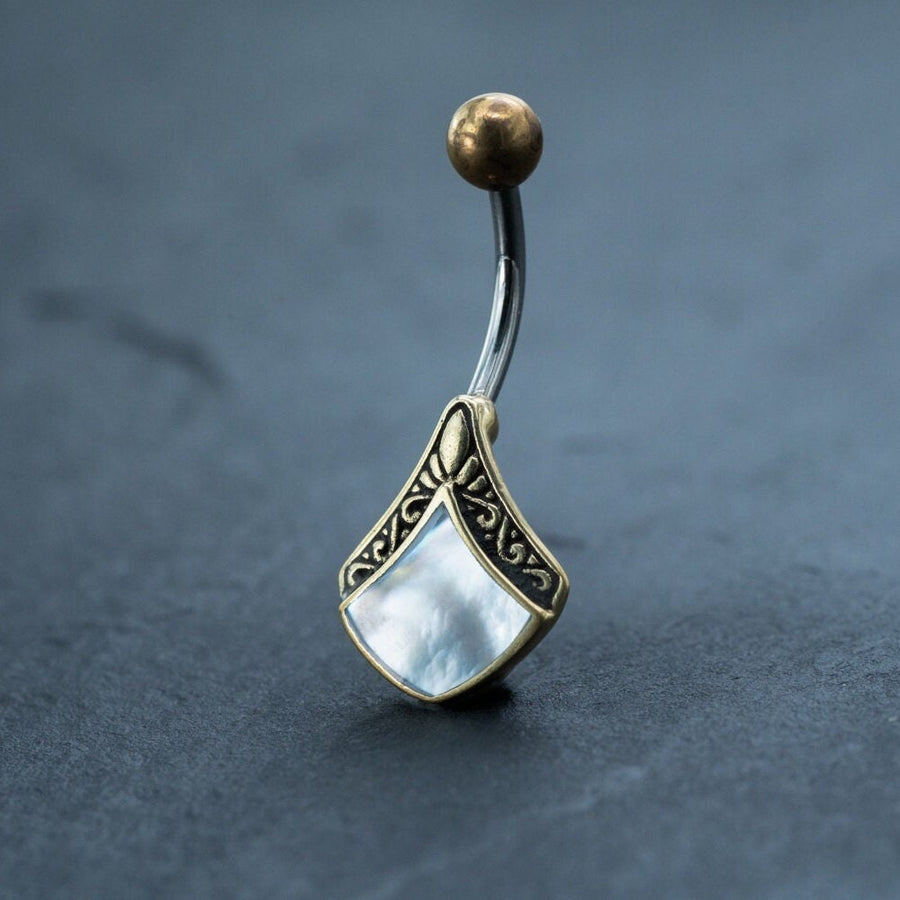 The Kaya Movement's Mother of Pearl Belly Ring - Boho 9mm Navel Bar is showcased. This diamond-shaped belly button jewelry features a curved metal barbell with a spherical gold-colored top. Its pendant includes an intricate gold-colored pattern and mother-of-pearl inlay, offering an elegant, boho look perfect for any navel piercing.