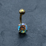 Introducing the Kaya Movement 18K Gold Navel Ring, a Crystal Belly Barbell that showcases two round, multi-colored opal-like gems. The smaller gem graces the top, while the larger gem adorns the bottom of this elegant piece of hypoallergenic body jewelry. Perfect for sensitive skin, this discreet piercing is shown against a dark gray, textured background.
