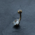 A close-up image of the Kaya Movement 18K Gold Navel Ring, Crystal & CZ Belly Barbell from their hypoallergenic body jewelry collection, features a curved barbell design with a small round clear gem at one end and a larger oval cubic zirconia accent securely set at the other, placed on a dark textured surface.
