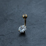 Depicted on a dark surface is the 18K Gold Navel Ring by Kaya Movement, featuring a curved barbell design. This hypoallergenic, allergy-friendly body jewelry showcases two clear, round-cut gemstones: a larger one at the bottom and a smaller one at the top.