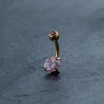 A gold curved barbell belly button ring with two pink crystals, one small and round at the top and a larger one oval-shaped at the bottom, is displayed on a dark, textured background. This stunning 18K Gold Navel Ring from Kaya Movement is hypoallergenic and perfect for those with sensitive skin.