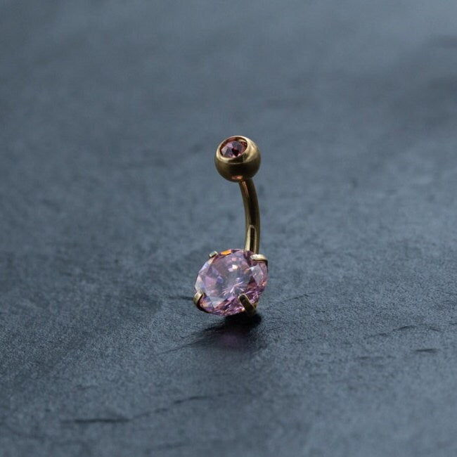 A gold curved barbell belly button ring with two pink crystals, one small and round at the top and a larger one oval-shaped at the bottom, is displayed on a dark, textured background. This stunning 18K Gold Navel Ring from Kaya Movement is hypoallergenic and perfect for those with sensitive skin.
