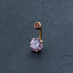 Experience the elegance of Kaya Movement's 18K Gold Navel Ring, featuring an exquisite pink crystal belly barbell. This hypoallergenic and discreet piercing showcases a large round pink gemstone at one end and a smaller pink gemstone at the other, set against a dark gray textured background. Perfectly eye-catching and delicate, this allergy-friendly body jewelry is ideal for those seeking a feminine design in their belly barbell collection.