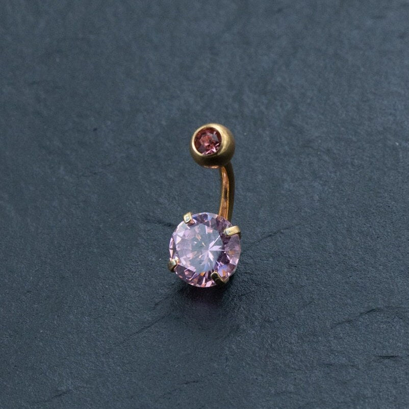 Experience the elegance of Kaya Movement's 18K Gold Navel Ring, featuring an exquisite pink crystal belly barbell. This hypoallergenic and discreet piercing showcases a large round pink gemstone at one end and a smaller pink gemstone at the other, set against a dark gray textured background. Perfectly eye-catching and delicate, this allergy-friendly body jewelry is ideal for those seeking a feminine design in their belly barbell collection.