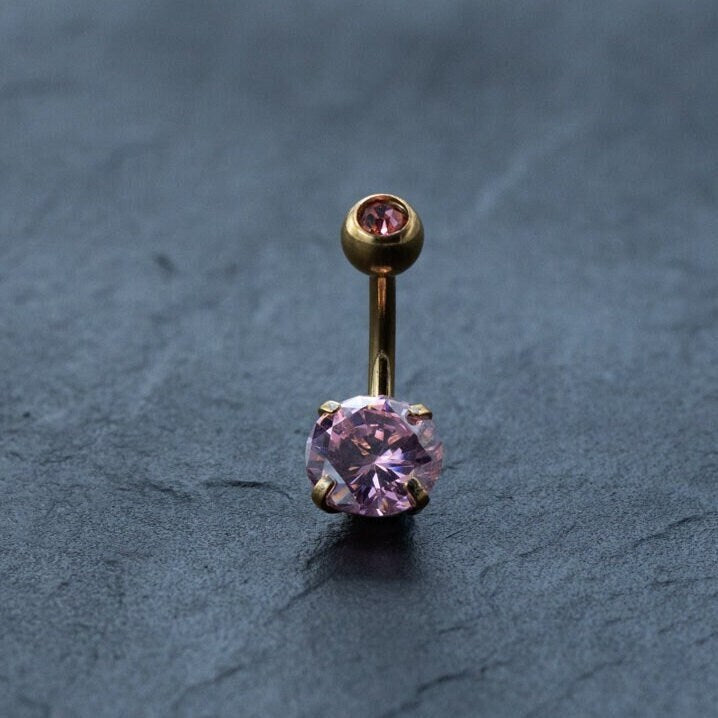 The Kaya Movement 18K Gold Navel Ring, known for its elegance and comfort, features two pink gemstones—a larger one at the bottom and a smaller one embedded in the top ball—set against a dark textured surface. This hypoallergenic belly barbell is perfect for those seeking discreet, allergy-friendly body jewelry.
