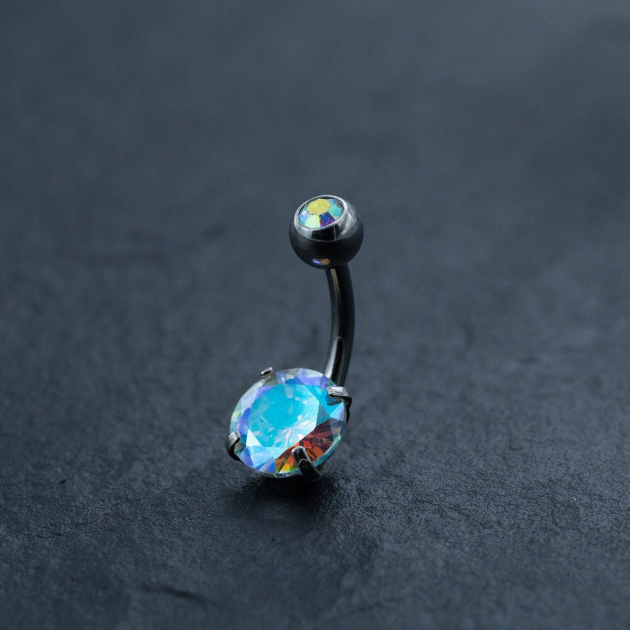 A black metal belly button ring from Kaya Movement rests on a dark textured surface. This Discreet Allergy-Friendly Crystal Belly Barbell is crafted from surgical silver for an allergy-friendly experience and features two iridescent zircon gems. The larger oval gem at the base and the smaller round gem at the top display a dazzling mix of blue, green, and purple hues, giving them a holographic appearance.