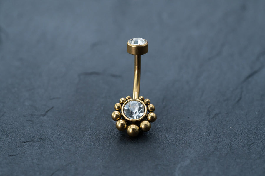 The Kaya Movement 18K Gold Navel Ring, named Crystal & CZ Belly Barbell, features clear cubic zirconia accents on each end. The larger gemstone is encircled by small gold beads in a circular pattern, adding an elegant touch. This hypoallergenic and discreet belly barbell is beautifully displayed on a dark, textured surface.