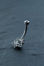 The Kaya Movement Hypoallergenic Crystal Zircon Belly Ring - Discreet CZ Navel Piercing is a silver hypoallergenic piece adorned with an intricately designed flower featuring petal-like structures and heart-shaped cutouts, each petal surrounding a clear gemstone at the center. Another small gemstone caps the end of the curved surgical steel barbell. The jewelry rests on a dark slate background.
