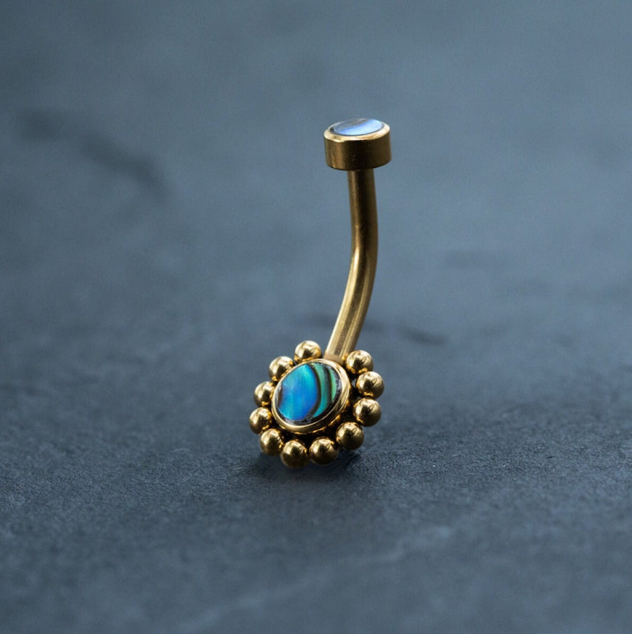 A stunning 18k navel piercing from Kaya Movement, this gold belly button ring features a curved bar with a circular abalone shell surrounded by delicate gold beads. An additional small, flat stud graces the top. The background is a smooth, dark surface showcasing this exquisite piece of boho-chic style and hypoallergenic body jewelry.