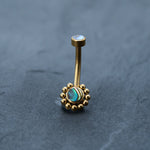 An 18k gold navel ring from Kaya Movement, part of their boho-chic collection, is exhibited on a dark gray surface. This beautiful piece of hypoallergenic belly jewelry has a curved barbell design, featuring a small opal-like stone at the top and an abalone shell in an intricate sunburst-like setting at the bottom.
