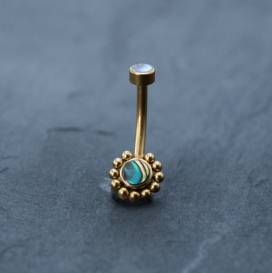 An 18k gold navel ring from Kaya Movement, part of their boho-chic collection, is exhibited on a dark gray surface. This beautiful piece of hypoallergenic belly jewelry has a curved barbell design, featuring a small opal-like stone at the top and an abalone shell in an intricate sunburst-like setting at the bottom.