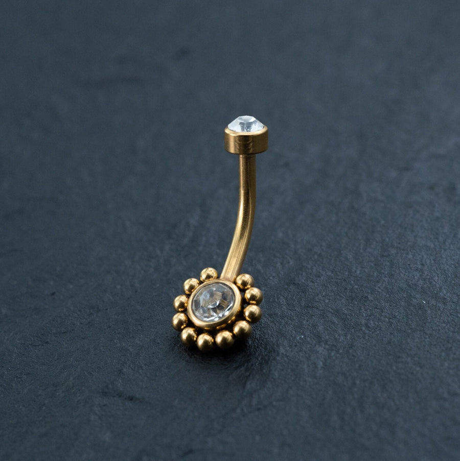A gold curved belly button ring from Kaya Movement is displayed on a dark, textured surface. The 18K Gold Navel Ring, named Crystal & CZ Belly Barbell, showcases a flower-shaped design at one end with a circular crystal center surrounded by small gold beads and cubic zirconia accents. At the other end, it features a single clear crystal set in gold. This hypoallergenic and allergy-friendly body jewelry ensures discreet and comfortable piercing wear.