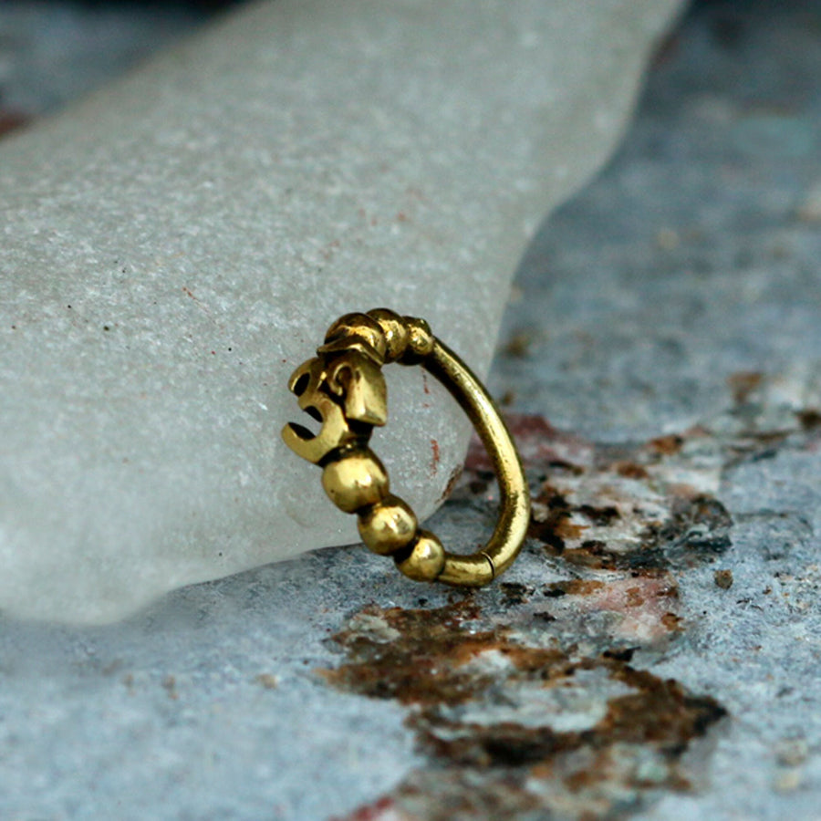 The Lotus Flower Helix Hoop from Kaya Movement, a seamless gold septum jewelry piece, sits delicately on a textured surface. Featuring an elegant "Om" symbol and adorned with intricate bead-like details, it evokes the traditional charm of Indian nose rings. The background showcases rough, earthy tones with a white, translucent object partially visible.