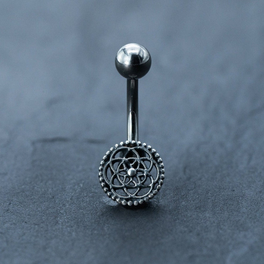 Introducing the Silver Mandala Belly Button Rings by Kaya Movement—a stunning surgical steel navel piercing boasting a 9mm curved barbell with an intricate flower-like mandala design at the center. Adorned with delicate bead details around the edges, this boho belly ring is perfect for navel piercings and stands out beautifully against a textured dark gray surface.