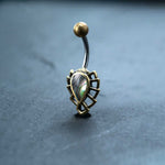 A close-up of the Kaya Movement Abalone Shell Belly Button Ring is shown on a flat surface. This 9mm Bananabell features iridescent colors set in a geometric, gold-toned frame with a small gold ball at the top, crafted from surgical steel. The background is a smooth, dark gray surface.