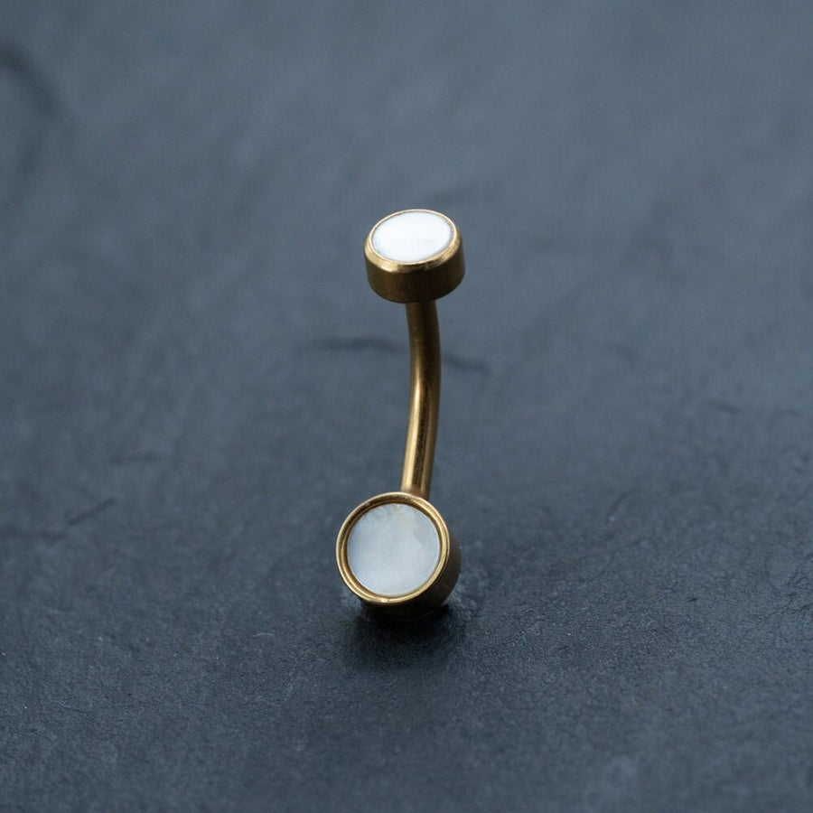 A curved, gold-tone barbell piercing with round white MOP shell stones set in both ends, displayed on a dark, textured surface. The minimalist design gives this Kaya Movement 18k Navel Piercing & Gold Navel Ring a modern, elegant look.