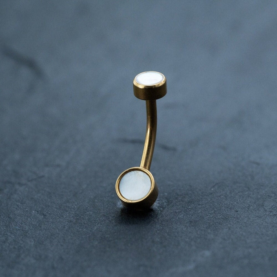 An 18k gold navel ring from Kaya Movement, adorned with round white MOP shell stones on each end, is placed vertically against a dark gray textured background.